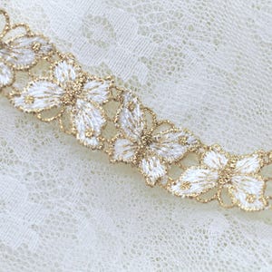 May include: A decorative trim featuring a repeating pattern of white embroidered butterflies with gold metallic thread outlines. The butterflies are connected in a continuous horizontal row, showcasing intricate stitching and a shimmering finish.