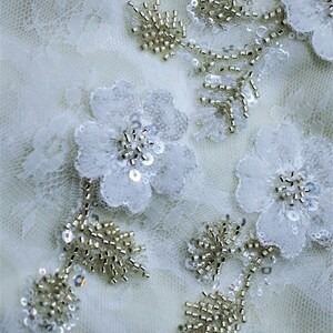 May include: White lace fabric with delicate floral embellishments and beaded accents. The flowers are made of white fabric and have a pearl-like sheen. The beads are small and gold in color.