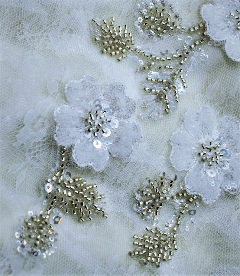 May include: White lace fabric with delicate floral embellishments and beaded accents. The flowers are made of white fabric and have a pearl-like sheen. The beads are small and gold in color.