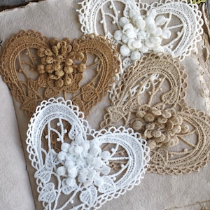 May include: Four heart-shaped lace appliqués in white and tan colors. Each piece features an intricate openwork pattern with a cluster of small, raised circular elements at the center. The lace hearts are arranged in a overlapping pattern.