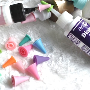 May include: A collection of small, conical, multi-colored plastic precision tips for glue bottles, including shades of pink, green, blue, orange, and purple, scattered alongside two white plastic squeeze bottles labeled Art Glitter and Diamond Glaze.