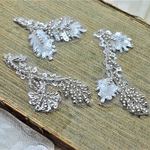 May include: Three white beaded floral appliques with silver sequins. The appliques are on a brown textured surface.