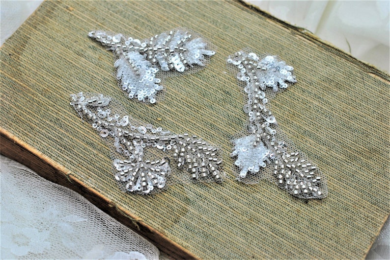 May include: Three white beaded floral appliques with silver sequins. The appliques are on a brown textured surface.