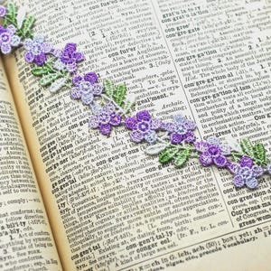 May include: A decorative lace trim featuring a repeating pattern of purple and light blue flowers with green leaves, laid diagonally across the open pages of a vintage dictionary with visible printed text columns.