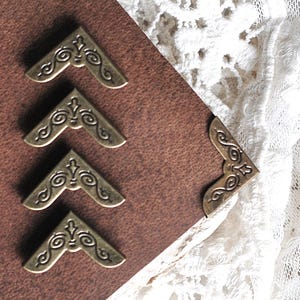 May include: Close-up of antique brass-colored corner protectors with ornate scrollwork, arranged on a brown leather surface. A piece of white lace is in the background. These decorative metal corners are for scrapbooking or bookbinding.
