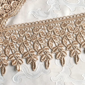 May include: A metallic gold lace trim featuring a repeating leaf pattern with a scalloped edge. The embroidery consists of dense, raised stitching forming individual leaves connected by a fine mesh structure, creating a decorative border.