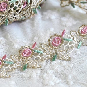 May include: A decorative lace trim featuring a repeating pattern of embroidered pink roses with small green leaves. The floral design is outlined in a metallic gold thread against a cream-colored, open-work mesh background with scalloped edges.