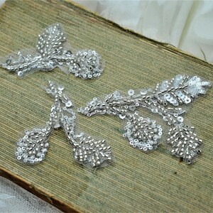 May include: Three silver beaded and sequined floral appliques on a green fabric background.