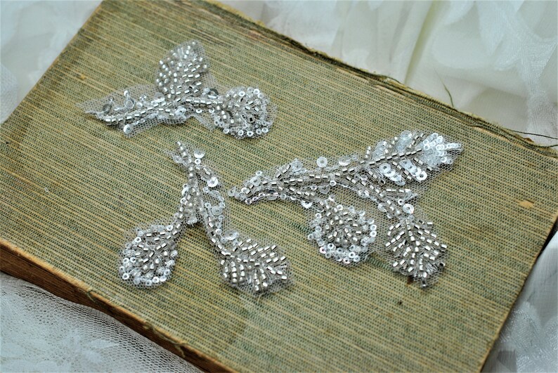 May include: Three silver beaded and sequined floral appliques on a green fabric background.