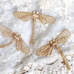 May include: Three delicate gold embroidered dragonfly appliques on a white lace background.
