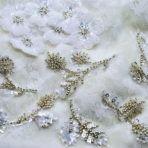 May include: White lace fabric with delicate floral and beaded embellishments. The fabric features a cluster of white flowers with pearl-like beads, as well as several beaded vines with small, round beads.