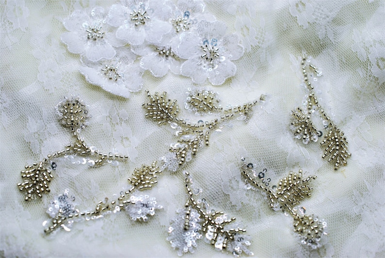 May include: White lace fabric with delicate floral and beaded embellishments. The fabric features a cluster of white flowers with pearl-like beads, as well as several beaded vines with small, round beads.