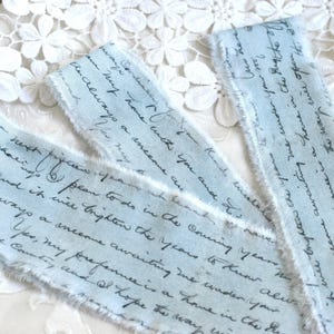May include: Three strips of light blue fabric ribbon featuring a dark blue cursive script print. Each ribbon has frayed, raw edges along the sides. The text on the fabric is legible and repeats across the surface of the material.