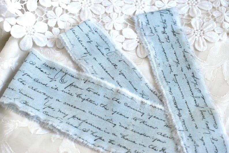 May include: Three strips of light blue fabric ribbon featuring a dark blue cursive script print. Each ribbon has frayed, raw edges along the sides. The text on the fabric is legible and repeats across the surface of the material.