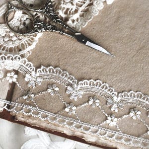 May include: A pair of silver-toned metal scissors with ornate handles resting on a piece of white lace trim featuring a floral pattern. The lace is positioned over a textured, light brown paper surface, creating a layered composition of materials.