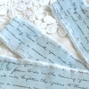 May include: Three strips of light blue fabric with frayed edges, featuring dark gray cursive handwriting printed across the surface. The text includes phrases such as 'always a welcome' and 'brighten the years to know'. The fabric has a soft texture.