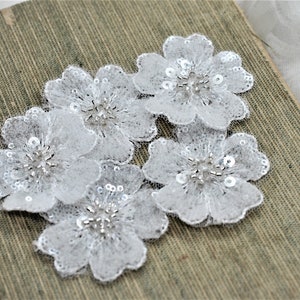 May include: Four white lace flower appliques with silver sequins and beads. The appliques are on a brown textured surface.