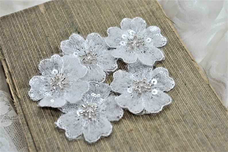 May include: Four white lace flower appliques with silver sequins and beads. The appliques are on a brown textured surface.