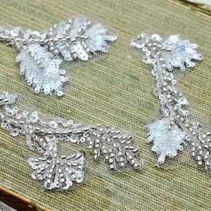 May include: Three white beaded floral appliques with silver sequins. The appliques are on a green fabric background.