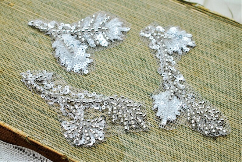 May include: Three white beaded floral appliques with silver sequins. The appliques are on a green fabric background.