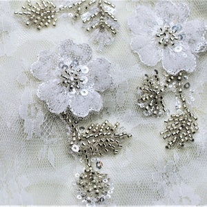 May include: White lace fabric with silver beaded floral embellishments. The fabric has a delicate, intricate pattern.