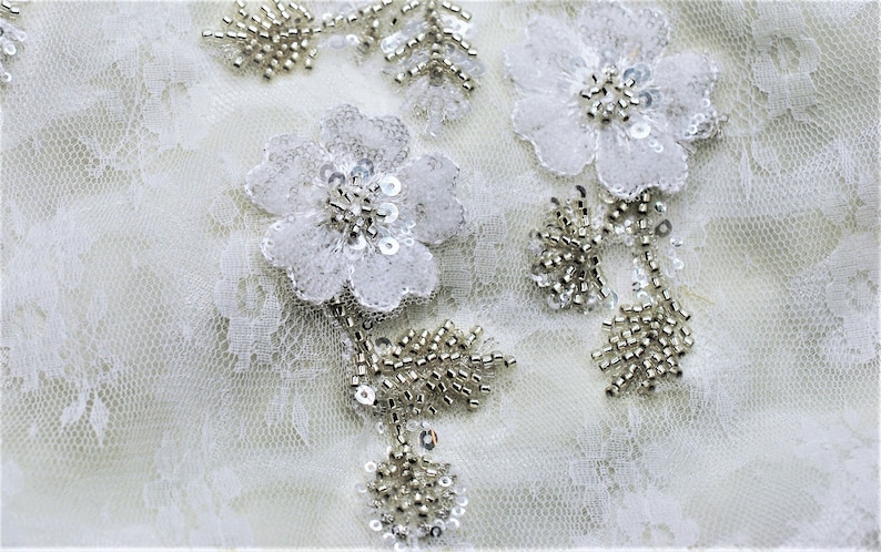May include: White lace fabric with silver beaded floral embellishments. The fabric has a delicate, intricate pattern.