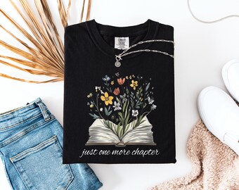 Peut inclure: T-shirt noir avec un motif floral et un livre ouvert. Le texte "just one more chapter" est imprimé sous le livre.
