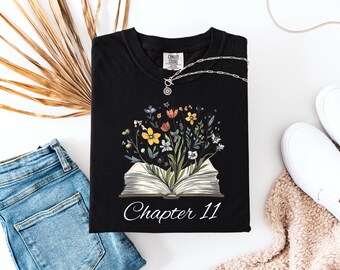 Peut inclure: T-shirt noir avec un motif floral émergeant d'un livre ouvert, avec le texte "Chapter 11" en blanc. Un collier argenté avec un pendentif est posé sur le t-shirt. Le t-shirt est accompagné d'un jean bleu et de baskets blanches.