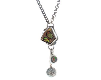 May include: A silver chain necklace featuring a pendant with a large, irregular, dark-toned stone with green and orange iridescent flecks, a smaller triangular stone inlay, and a round, etched silver bead dangling at the bottom of the piece.