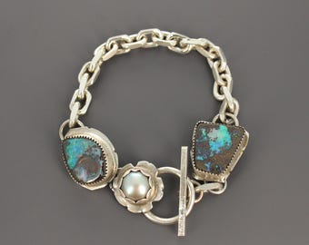 May include: A silver chain link bracelet featuring two irregular, dark-toned stones with vibrant blue and green iridescent flecks, and a central round white pearl set within a scalloped silver flower-shaped frame, secured by a silver toggle clasp.