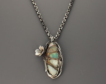 May include: A silver pendant necklace featuring an oval-shaped setting with four inlaid iridescent opal stones. The metal frame has a dark, oxidized finish and includes a small, sculpted silver flower detail attached to the side of the pendant.