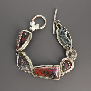Könnte beinhalten: Ein Silberarmband mit Knebelverschluss, bestehend aus fünf Gliedern mit geschichteten, mehrfarbigen Mustern in Rot, Grün, Schwarz, Weiß und Blau. Die Glieder sind in gezackte Silberfassungen eingefasst und durch Silberringe miteinander verbunden.