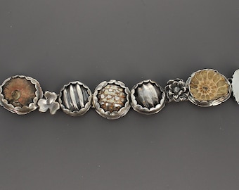 May include: A silver bracelet featuring a series of circular settings containing various fossils and textured stones, interspersed with small silver floral and leaf accents. The piece is secured with a toggle clasp consisting of a bar and a ring.