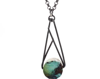 May include: A necklace featuring a dark, oxidized metal chain and a geometric pendant frame. The frame holds a round, polished turquoise stone with prominent dark brown matrix veining, suspended from a simple circular bail at the top.