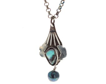 May include: A silver pendant necklace featuring a central iridescent stone with blue and green hues, set in a textured silver frame with radiating lines, suspended from a silver chain with a dark, round dangling bead at the bottom.