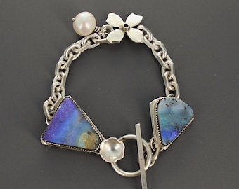 May include: A silver chain link bracelet featuring two triangular iridescent blue and purple opal-like stones in serrated settings, a single white pearl charm, a four-petaled flower charm, and a toggle clasp with a small flower-shaped accent.