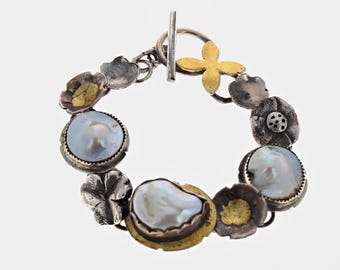 May include: A bracelet featuring three iridescent white pearls set in silver, interspersed with oxidized silver flower-shaped charms and gold-toned accents. The piece is secured with a silver toggle clasp featuring a bar and ring closure.