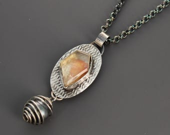 May include: A necklace featuring a dark oxidized silver chain and a pendant with an oval textured silver plate. The plate holds a faceted amber-colored stone, and a dark gray spherical bead is encased in a silver wire coil at the bottom.