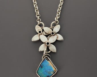 May include: A silver necklace featuring a pendant with three small, flat, five-petaled flowers arranged in a cluster. Suspended from the floral cluster is a single, irregular, kite-shaped stone with a vibrant blue and black iridescent surface.