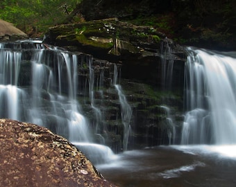 May include: A scenic waterfall cascades over layered rocks, creating a flowing curtain of water. The water is a bright white, contrasting with the dark rocks and surrounding green foliage. A large, textured rock is in the foreground.