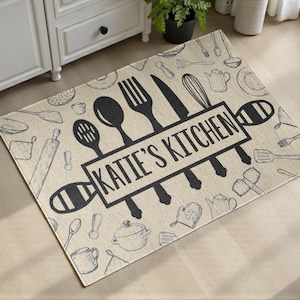 Può includere: Tappeto da cucina beige con un design di utensili in bianco e nero. Il tappeto presenta la scritta "KATIE'S KITCHEN" in una cornice rettangolare, con un motivo a matterello su ogni lato. A tema cucina.