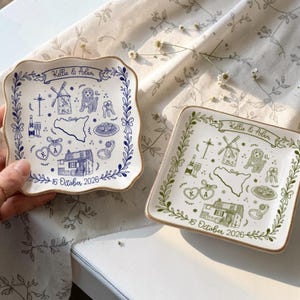 May include: Two square ceramic trinket dishes with scalloped edges and gold-painted rims. One dish features blue ink illustrations of a windmill, a dog, a house, and the text Kellie & Adam 16 October 2026. The other dish has identical green ink.