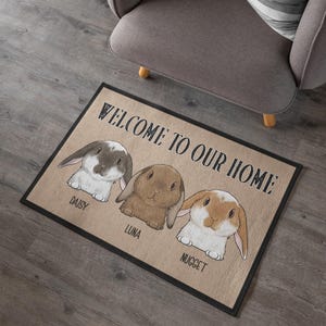 May include: Rectangular doormat with a black border and the text "WELCOME TO OUR HOME." Features illustrations of three rabbits named Daisy, Luna, and Nugget. The mat has a neutral color scheme, with the rabbits in shades of brown and white.
