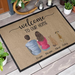 May include: A rectangular doormat with a black border and a tan background. The mat features the text "welcome TO OUR HOME" in black script. Below the text are illustrations of a couple and two pets, with names below them.