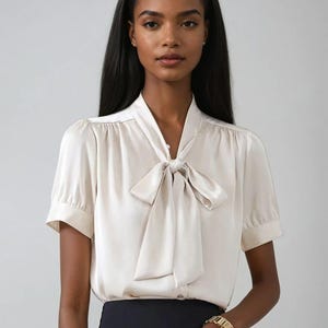 May include: Cream-coloured blouse with a necktie bow. The short-sleeved top has a classic design. The model is wearing a black skirt and a gold bracelet. The background is a solid light grey.