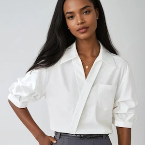 Può includere: Una camicia bianca a maniche lunghe con un esclusivo design a portafoglio e una tasca sul petto. La camicia è abbinata a una gonna a pieghe grigia e una cintura nera. È visibile una collana d'oro.