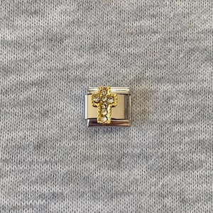 May include: A gold and silver charm featuring a cross design. The cross is adorned with small, clear gemstones. The charm is rectangular with a silver base and gold detailing. The background is a grey fabric.