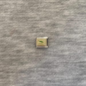 May include: A square modular link charm featuring a polished silver-tone metal frame and a cream-coloured enamel inlay. The centre of the charm displays a small, stylized green lizard graphic with a tiny red dot detail on its head.