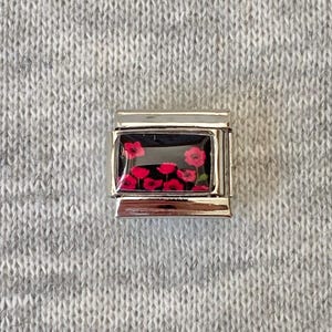 May include: A rectangular stainless steel charm link featuring a glossy black inlay decorated with a pattern of vibrant red poppy flowers. The charm has a polished metallic frame with horizontal bars at the top and bottom for attachment.