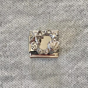 Op de afbeelding: Een vierkante, zilverkleurige modulaire bedel-schakel met een centrale uitsparing, omringd door een decoratieve opstelling van een zeester, een schelp en diverse kleine, heldere, ronde strass-steentjes in het metalen frame.